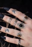 hand with occult tattoos and black nails, paired with sterling silver rings, in the middle a large black stone ring, surrounded by 2 golden brown tiger eye rings.