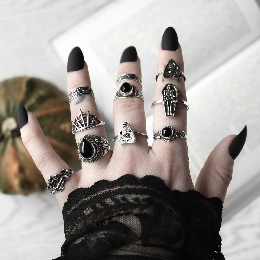 Hand with multiple Halloween themed sterling silver rings, symbols vary from spider webs, creepy crawlers to coffins, some rings are adorned with black Onyx stones, light background with an open book and a small pumpkin