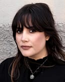 Raven-haired woman wears a recycled sterling silver necklace with intricate details and oxidised finish, the necklace is a skull with mushrooms on its side, enframed with a branch with leaves at the bottom