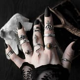 Hand with multiple sterling silver rings, 1 large ring in shape of a serpent with head slithering along the finger and tail pointing towards the arm, 1 ring with pentagram symbol, 2 rings with ocult inscriptions, 2 rings with golden brown Tiger eye stones, 1 ring in shape of 2 entwined snakes, dark background with occult symbols