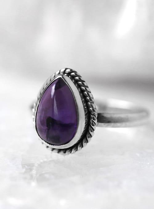 A sterling silver ring witha a drop shaped, deep purple Amethyst stone, sitting on a white crystal, white background