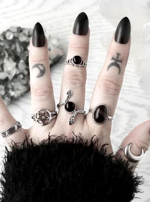 hand with occult finger tattoos and long black nails on white background showing multiple silver rings, from the left - 1 sacred geometry pattern silver ring, 1 pentgram ring in sterling silver, 1 snake ring with black onyx, 1 rutile ring in sterling silver and on the tumb 1 moon ring.