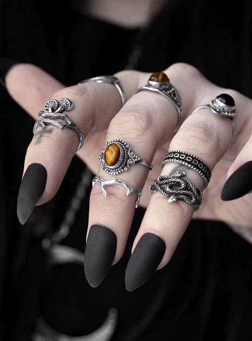 Close-up view of a hand with multiple sterling silver rings, 1 ring shaped like a branch with mushrooms, 1 ring with 2 entwined snakes, 1 ring with a genuine golden brown Tiger eye stone, dark background