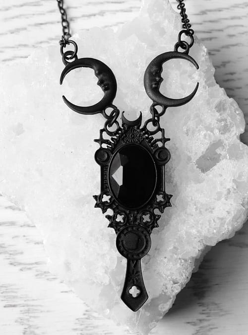 A black necklace laying on a crystal quartz stone. The necklace has a black stone in the middle which is surrounded by a frame with pagan symbols and crescent moons.