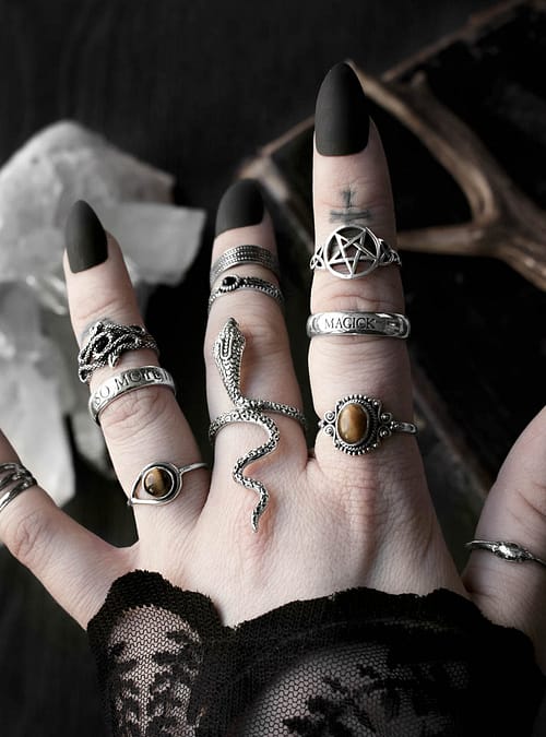 Hand with multiple sterling silver rings, 1 large ring in shape of a serpent with head slithering along the finger and tail pointing towards the arm, 1 ring with pentagram symbol, 2 rings with ocult inscriptions, 2 rings with golden brown Tiger eye stones, 1 ring in shape of 2 entwined snakes, dark background with occult symbols