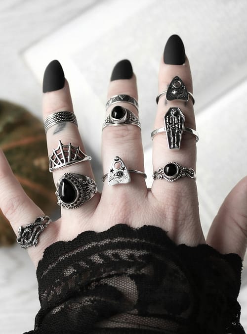 Hand with multiple Halloween themed sterling silver rings, symbols vary from spider webs, creepy crawlers to coffins, some rings are adorned with black Onyx stones, light background with an open book and a small pumpkin