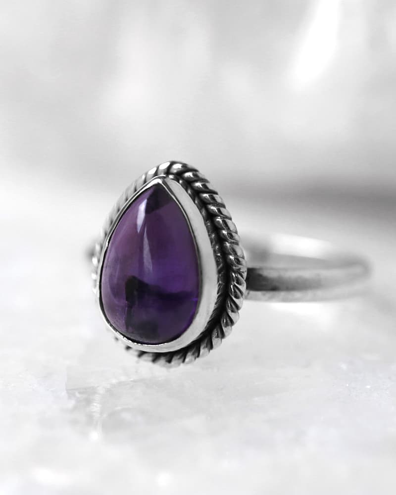 A sterling silver ring witha a drop shaped, deep purple Amethyst stone, sitting on a white crystal, white background