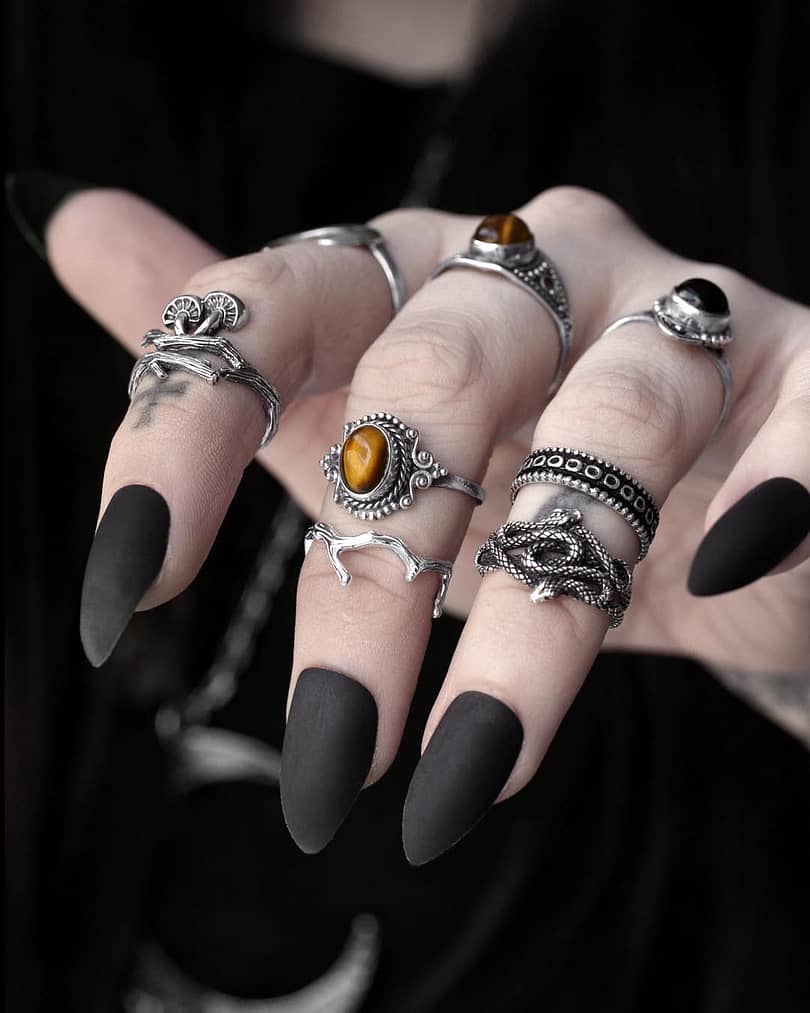 Close-up view of a hand with multiple sterling silver rings, 1 ring shaped like a branch with mushrooms, 1 ring with 2 entwined snakes, 1 ring with a genuine golden brown Tiger eye stone, dark background