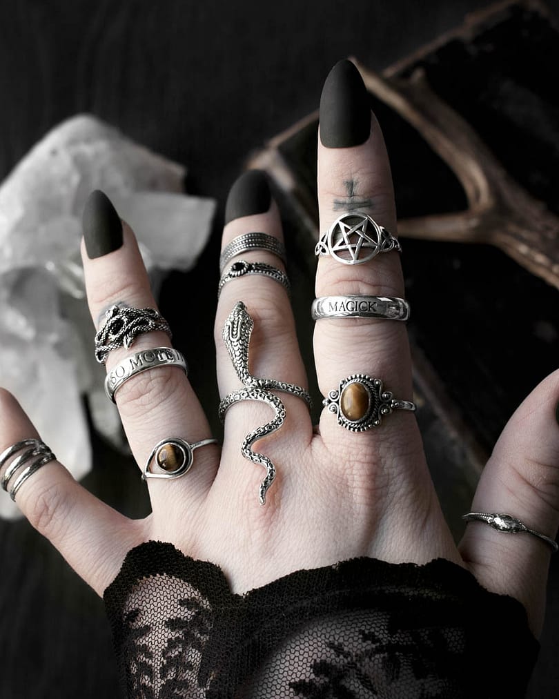 Hand with multiple sterling silver rings, 1 large ring in shape of a serpent with head slithering along the finger and tail pointing towards the arm, 1 ring with pentagram symbol, 2 rings with ocult inscriptions, 2 rings with golden brown Tiger eye stones, 1 ring in shape of 2 entwined snakes, dark background with occult symbols