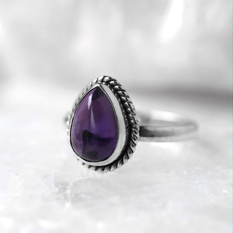 A sterling silver ring witha a drop shaped, deep purple Amethyst stone, sitting on a white crystal, white background