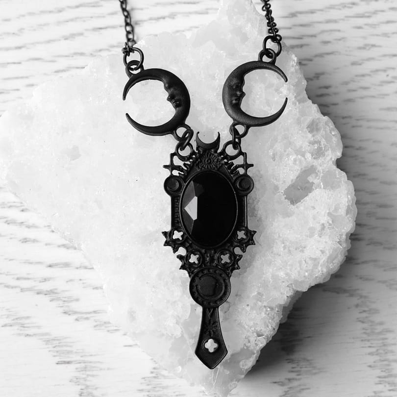 A black necklace laying on a crystal quartz stone. The necklace has a black stone in the middle which is surrounded by a frame with pagan symbols and crescent moons.